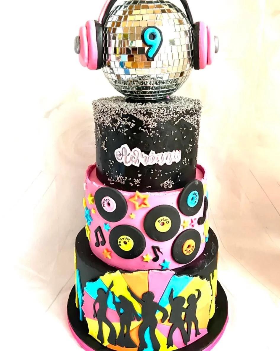 Three-tier disco themed cake with a large silver disco ball topper, pink headphones, vinyl records, and dancing silhouettes.