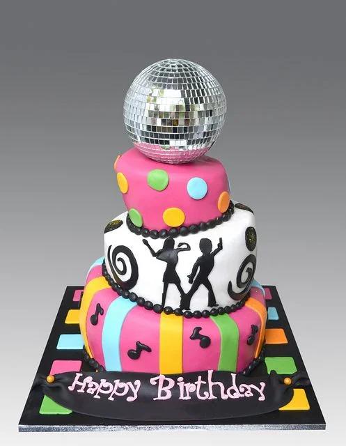 Three-tier colorful disco-themed birthday cake with a large silver disco ball topper, dancing silhouettes, and musical notes.