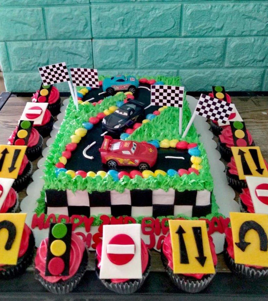 Rectangle racing car themed cake with Lightning McQueen and friends on a black track with green grass icing and checkered borders.