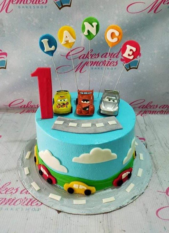 Light blue Disney Cars themed birthday cake with toy cars, balloon name toppers, and a red number one candle.