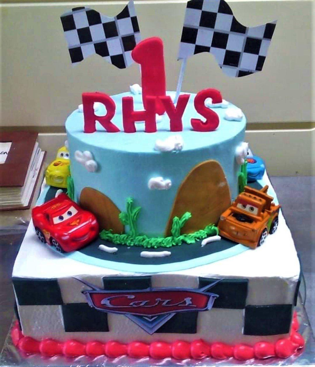 Two-tier Disney Cars themed birthday cake with blue sky top tier, checkered bottom tier, and toy car toppers including Lightning McQueen and Mater.