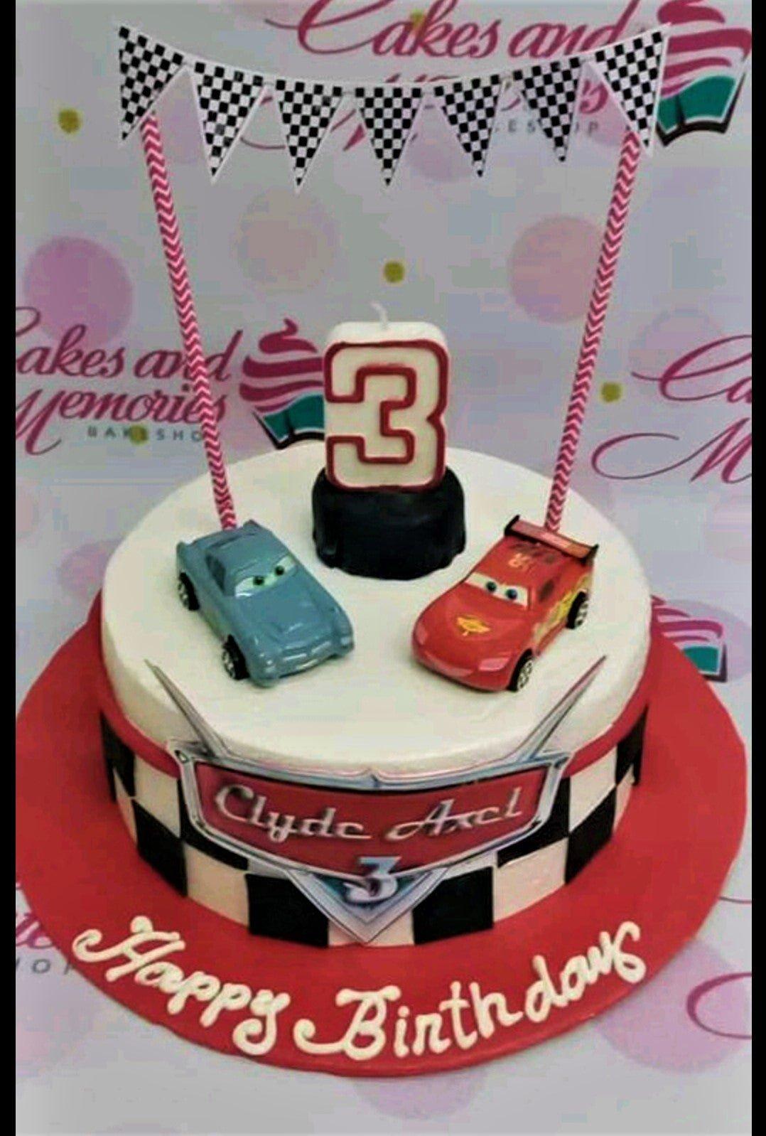 Disney Cars themed birthday cake with Lightning McQueen and Tow Mater toy cars, checkered flag banner, and a number 3 candle.