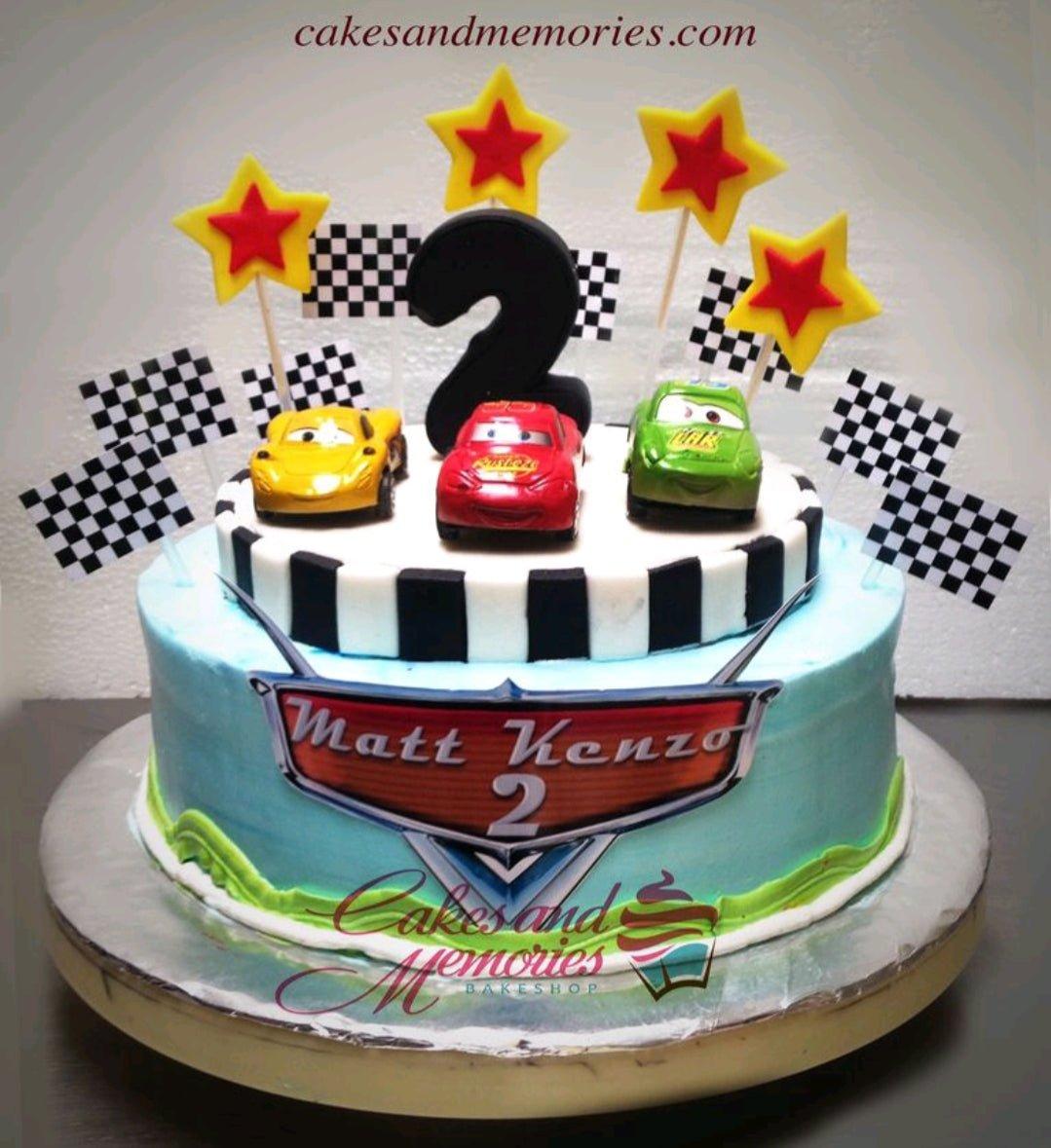 Disney Cars themed 2-tier birthday cake with Lightning McQueen, Mater, and Jackson Storm toy cars, checkered flags, and yellow stars.