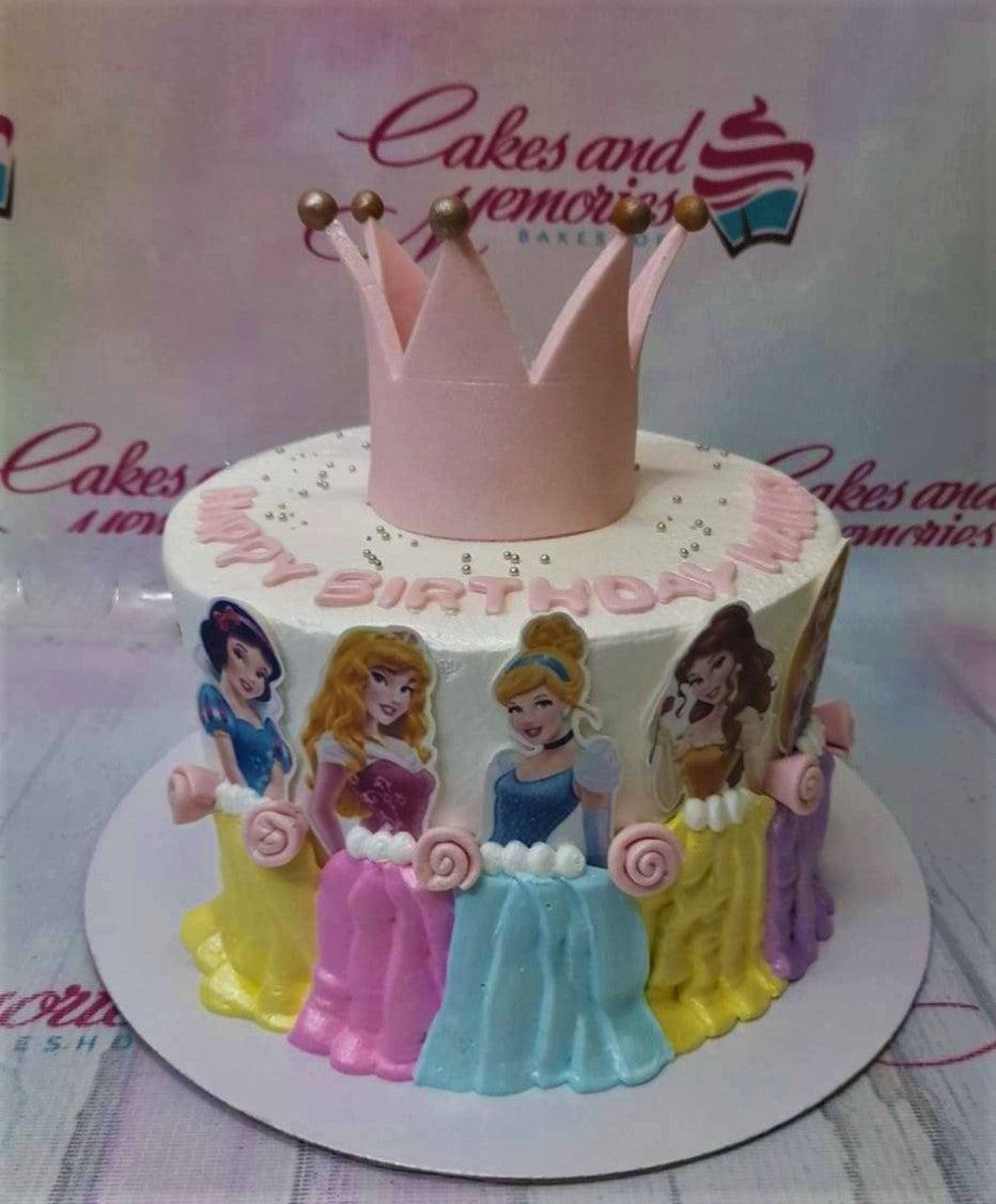 White 1-tier Disney Princess birthday cake with a pink crown topper and colorful icing dress designs for Cinderella, Belle, and Aurora.