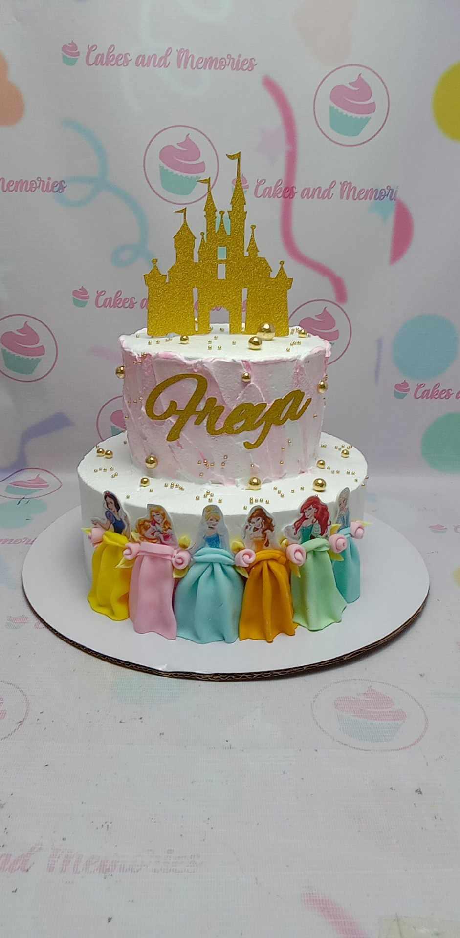Two-tier Disney Princess themed cake in pink and white with a gold castle topper and fondant princess figures.
