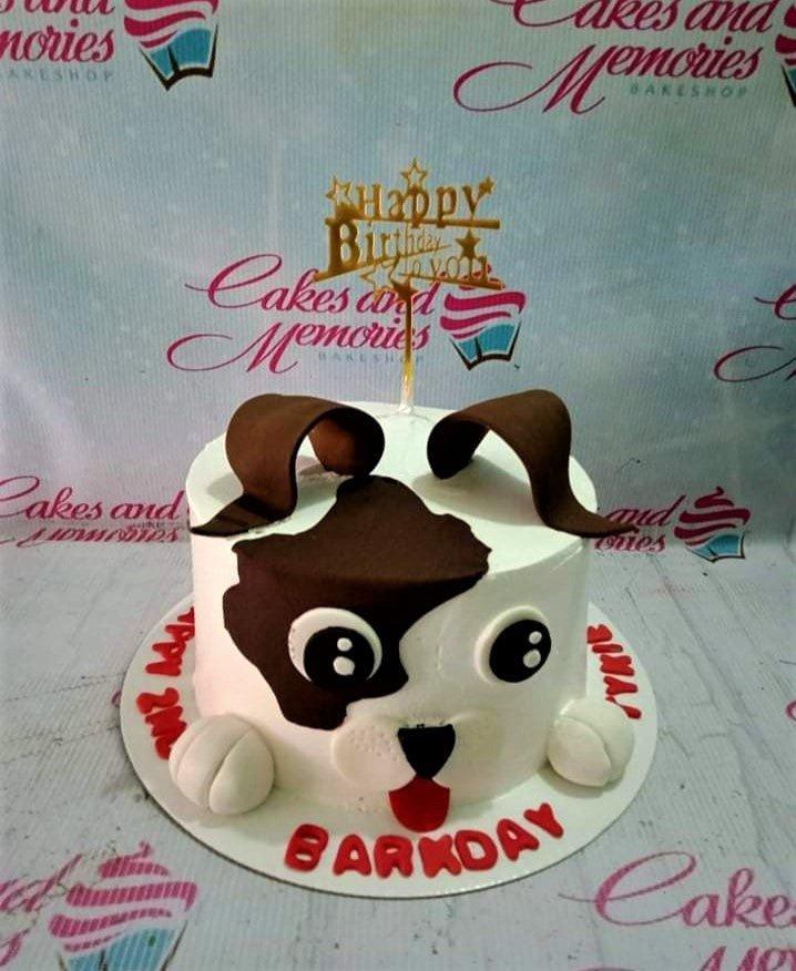 White dog-themed birthday cake with brown fondant ears, a spot over one eye, and a gold Happy Birthday topper.