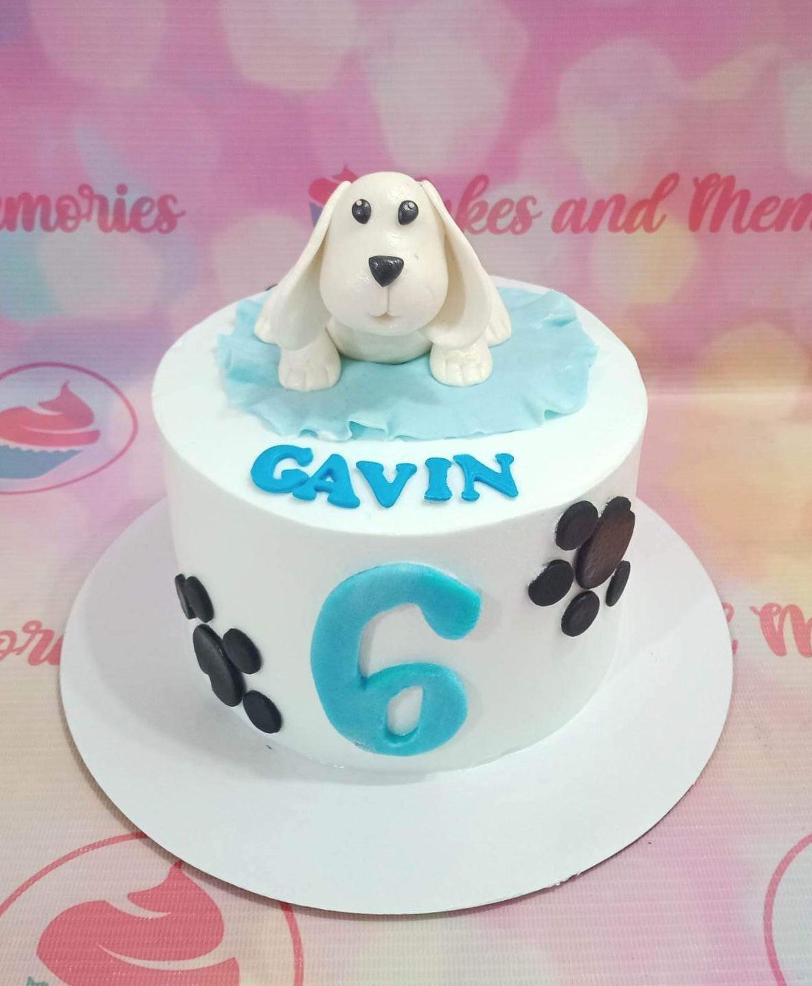 White round cake with a 3D fondant dog topper, blue name Gavin, blue number 6, and black paw prints on the side.