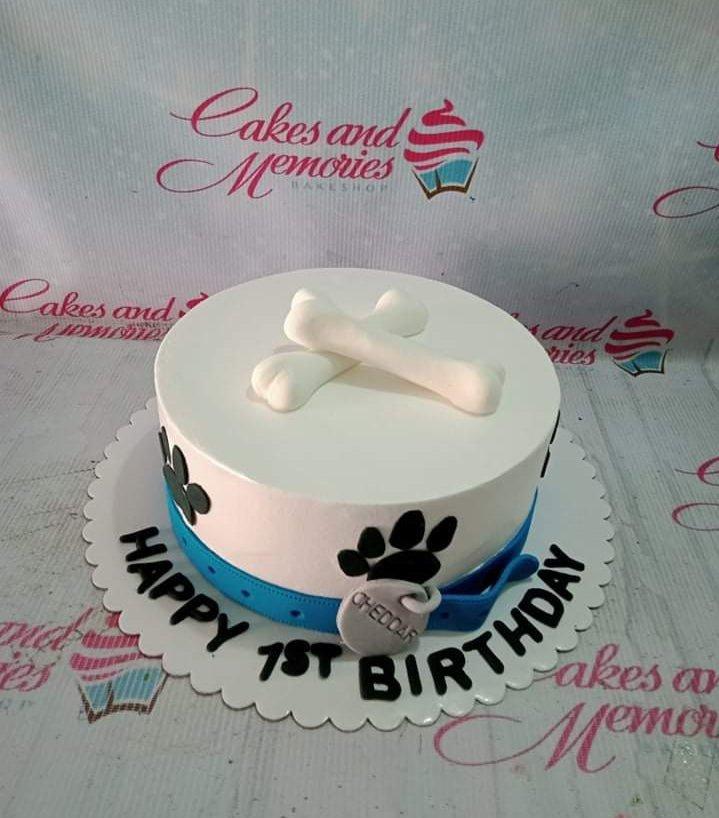 White dog-themed birthday cake with fondant bones on top, black paw prints on the side, and a blue collar with a name tag.