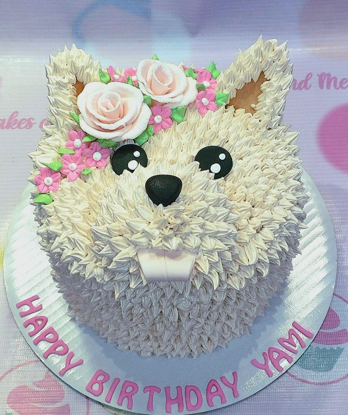 White dog-shaped cake with cream-colored icing fur, pink flowers, and a Happy Birthday Yami message on the base board.