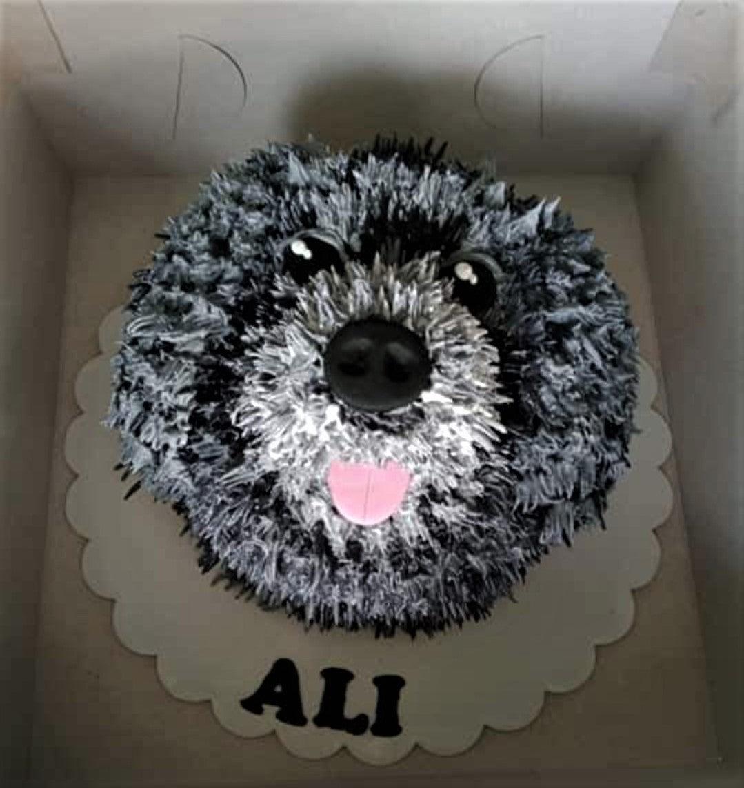 Black and white dog face cake with textured icing fur, large black nose, and pink tongue on a white scalloped board.