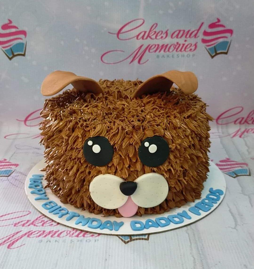 Brown shaggy dog face cake with floppy ears, large black eyes, and a pink tongue on a white board with a birthday message.
