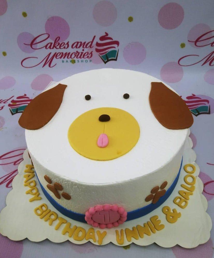 White dog face cake with brown ears, yellow muzzle, and pink tongue, featuring paw prints and a blue ribbon border.