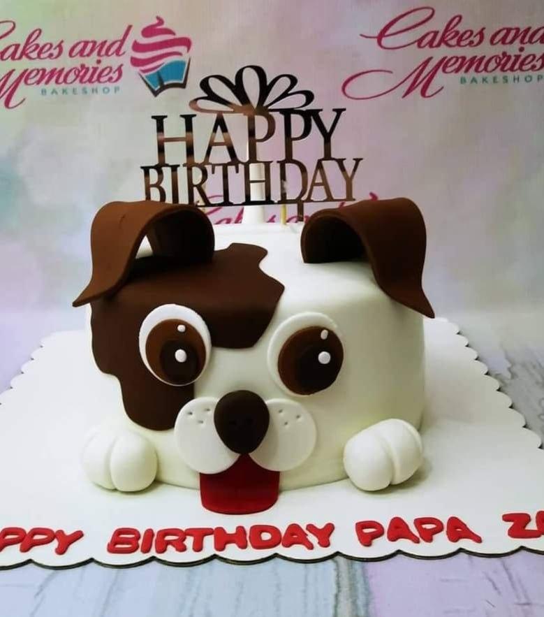 White fondant dog face cake with brown patches, floppy ears, and a red tongue, featuring a Happy Birthday topper.