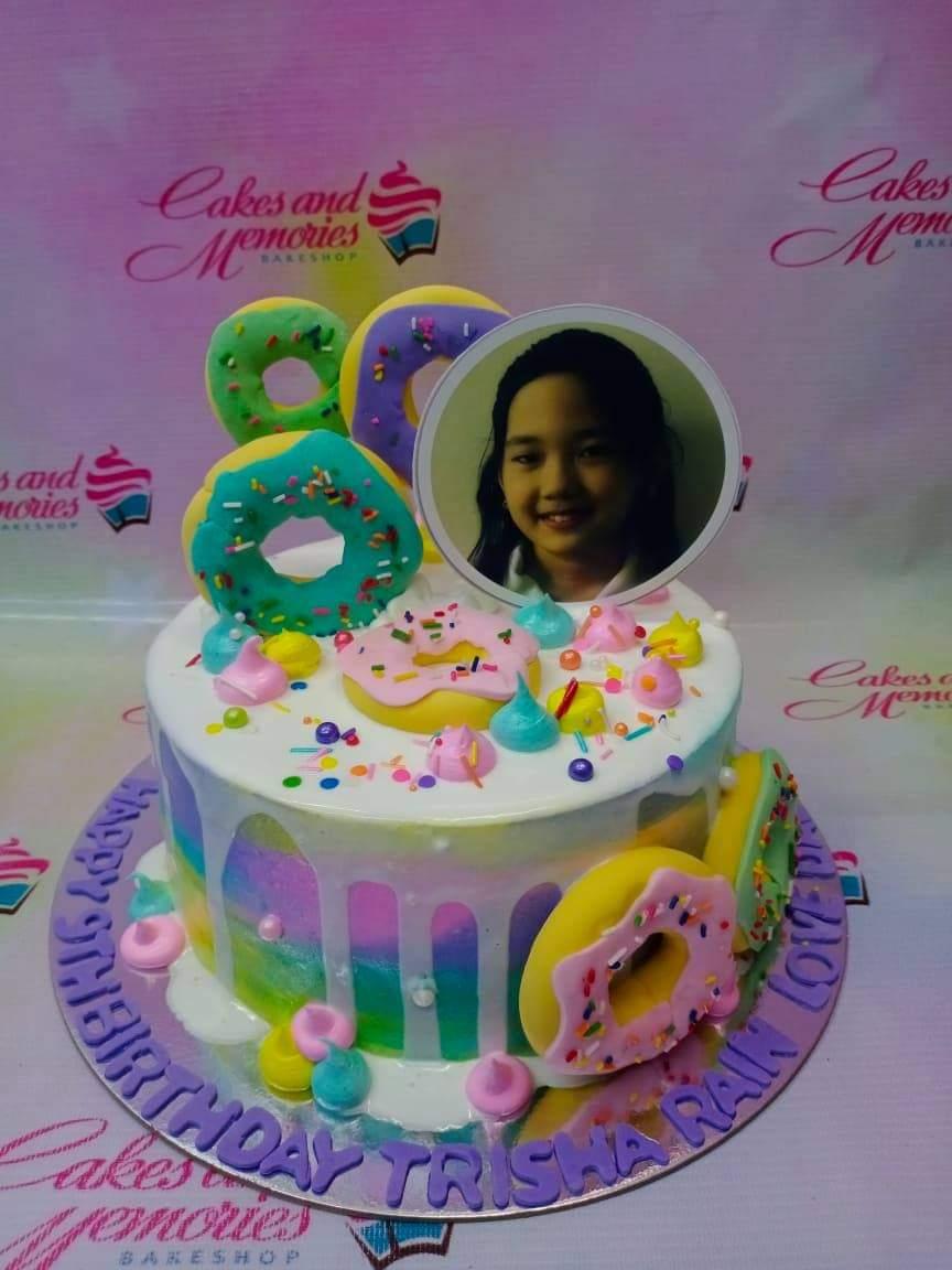 Colorful donut themed birthday cake with a photo printout topper, pastel icing drips, and fondant donut decorations.