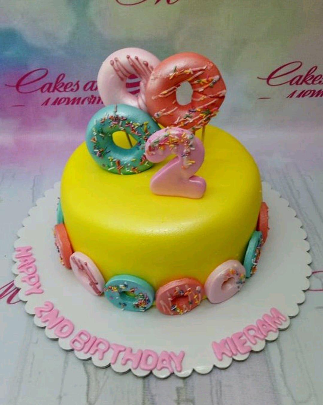 Yellow fondant cake decorated with colorful 3D fondant donuts and a pink number 2 topper for a 2nd birthday celebration.