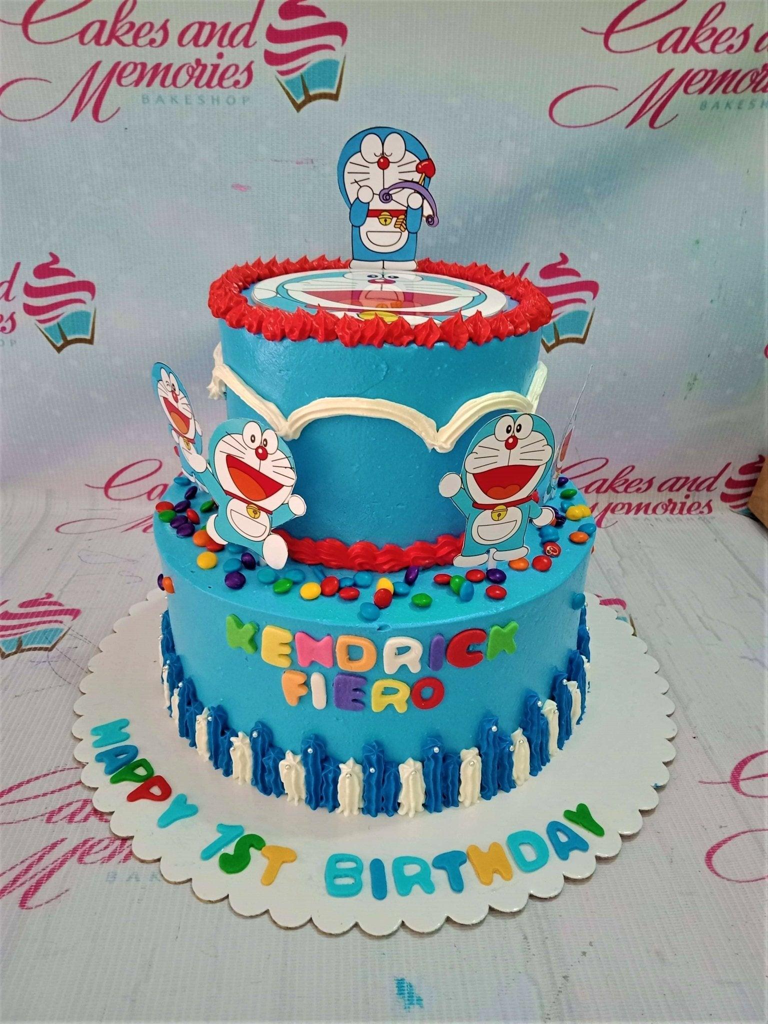 Two-tier blue Doraemon themed birthday cake with character printouts, colorful chocolate candies, and white and blue icing borders.