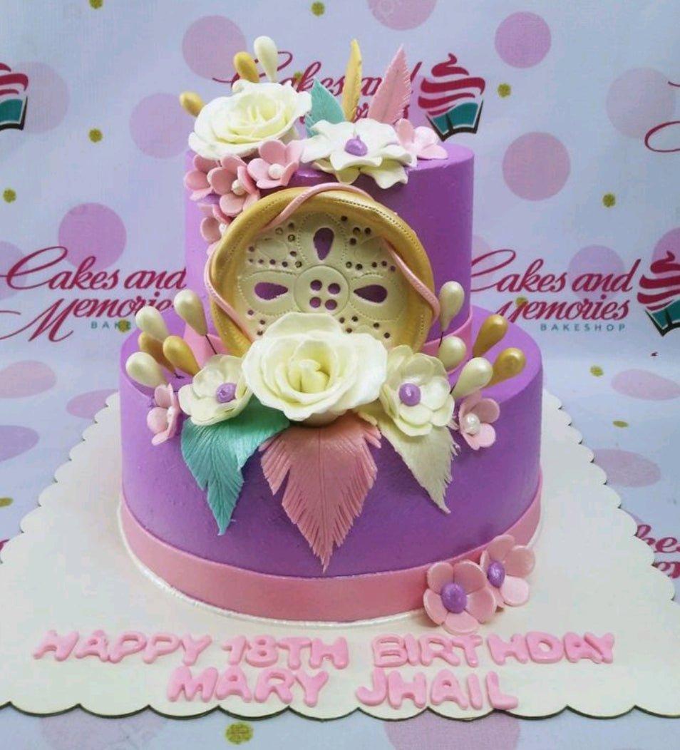 Two-tier purple fondant cake with a dreamcatcher topper, white and pink sugar flowers, and gold accents.