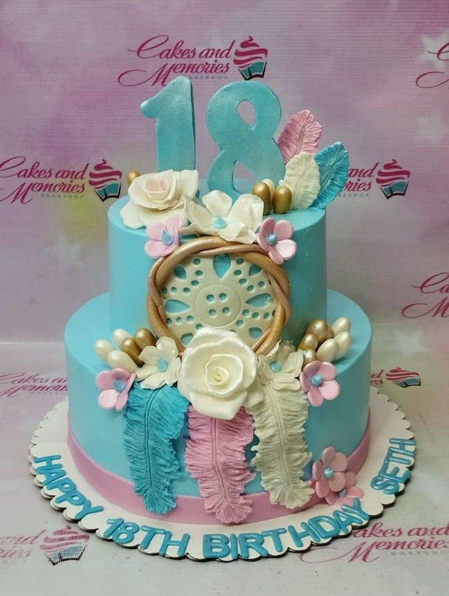 Two-tier light blue fondant dreamcatcher cake with pink and white flowers, feathers, and a large number 18 topper.