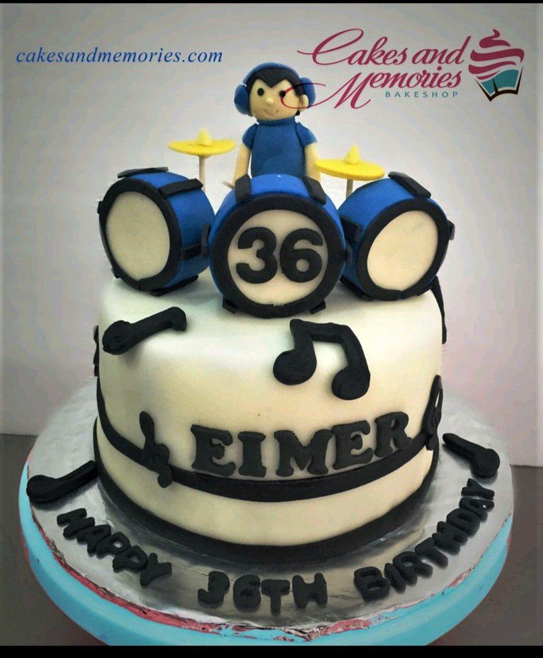 White fondant drum set birthday cake with a 3D drummer figure, blue drums, and black musical notes.