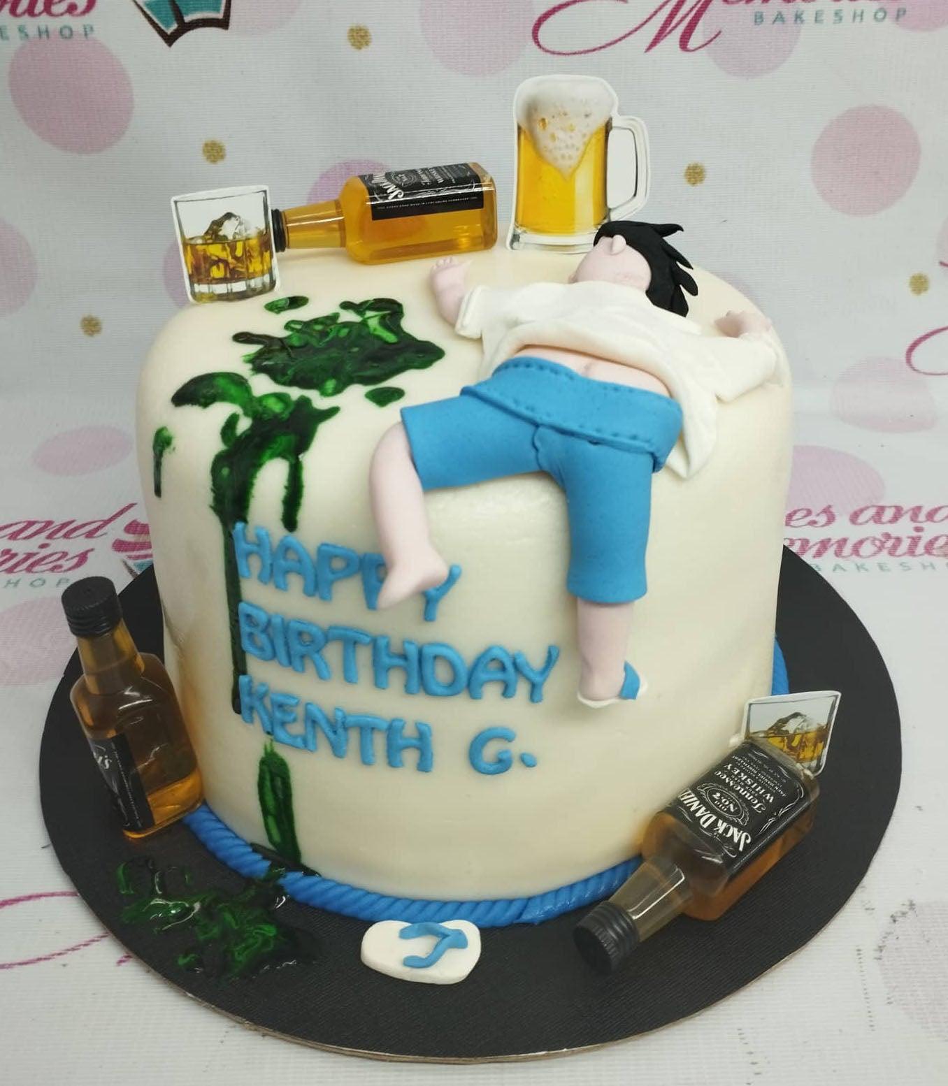 A humorous white fondant cake featuring a 3D edible figure of a man passed out next to spilled green liquid and miniature whiskey bottles.