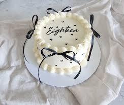 White minimalist vintage cake with black ribbon bows and piped icing borders featuring the word Eighteen in black script.