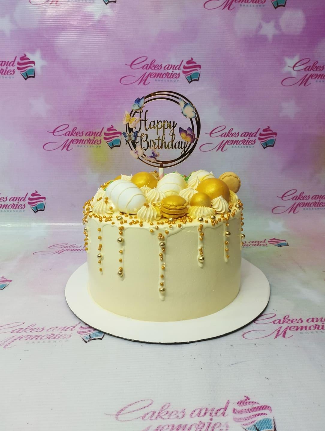 Elegant cream-colored birthday cake with gold drips, white and gold spheres, macarons, and a butterfly-themed Happy Birthday topper.