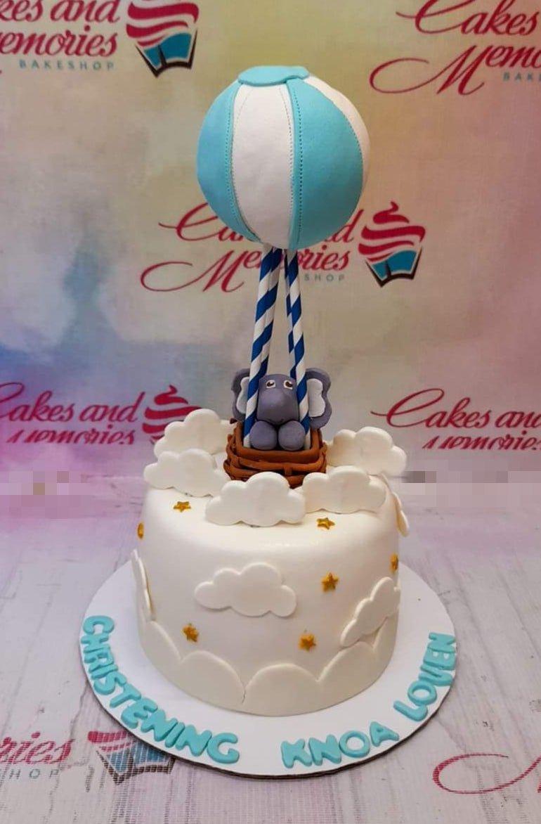 White fondant christening cake with a 3D elephant in a hot air balloon topper, white clouds, and gold stars.