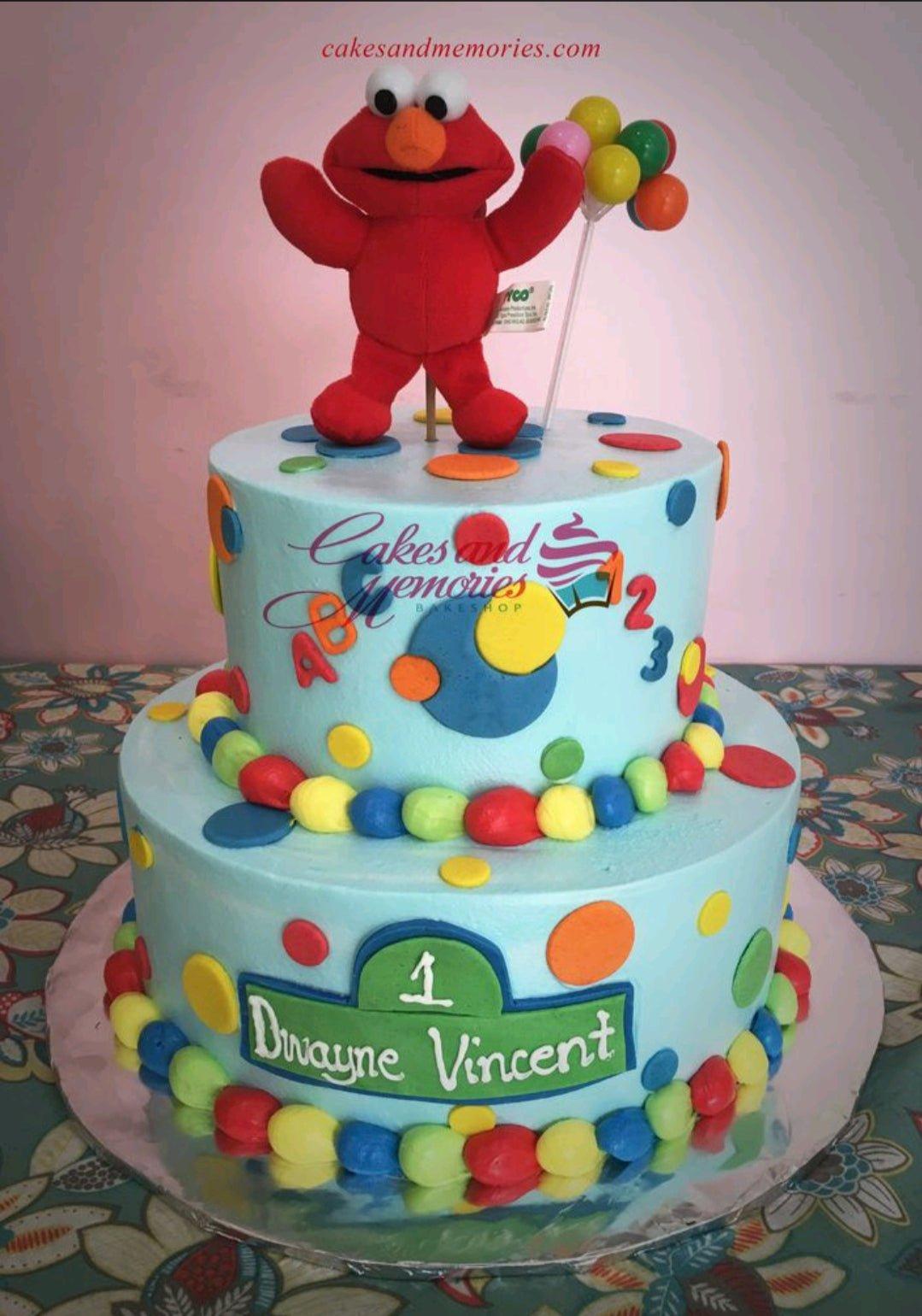 Two-tier light blue Elmo themed birthday cake with colorful fondant dots and a Sesame Street style name plaque.
