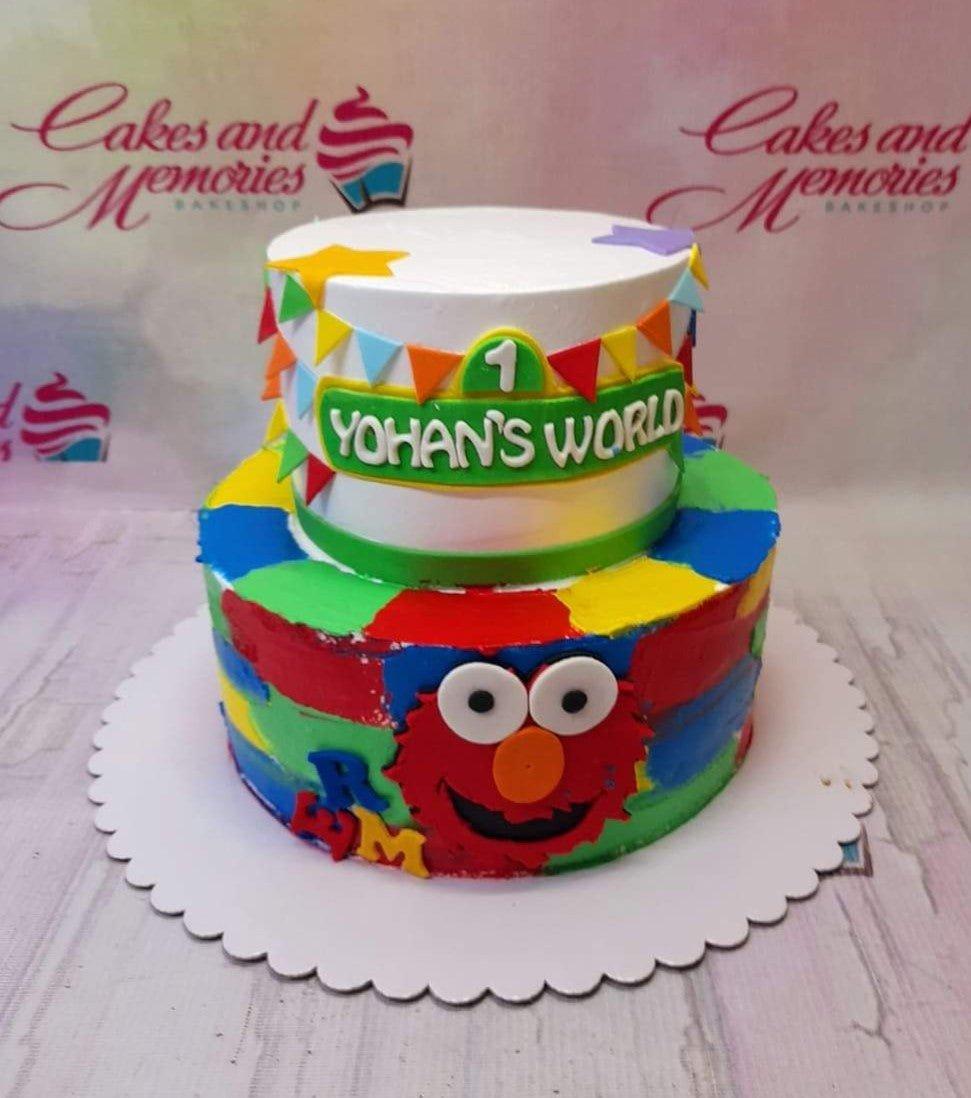 Two-tier Elmo and Sesame Street themed birthday cake with colorful icing patches and a custom name banner.