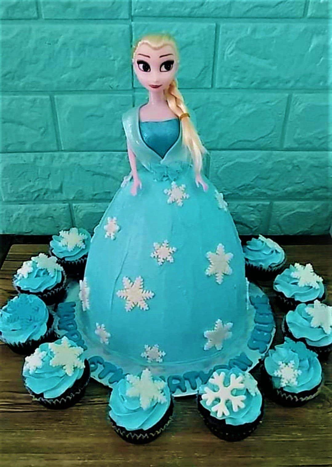 Elsa doll cake with a light blue icing dress decorated with white snowflakes and surrounded by matching cupcakes.