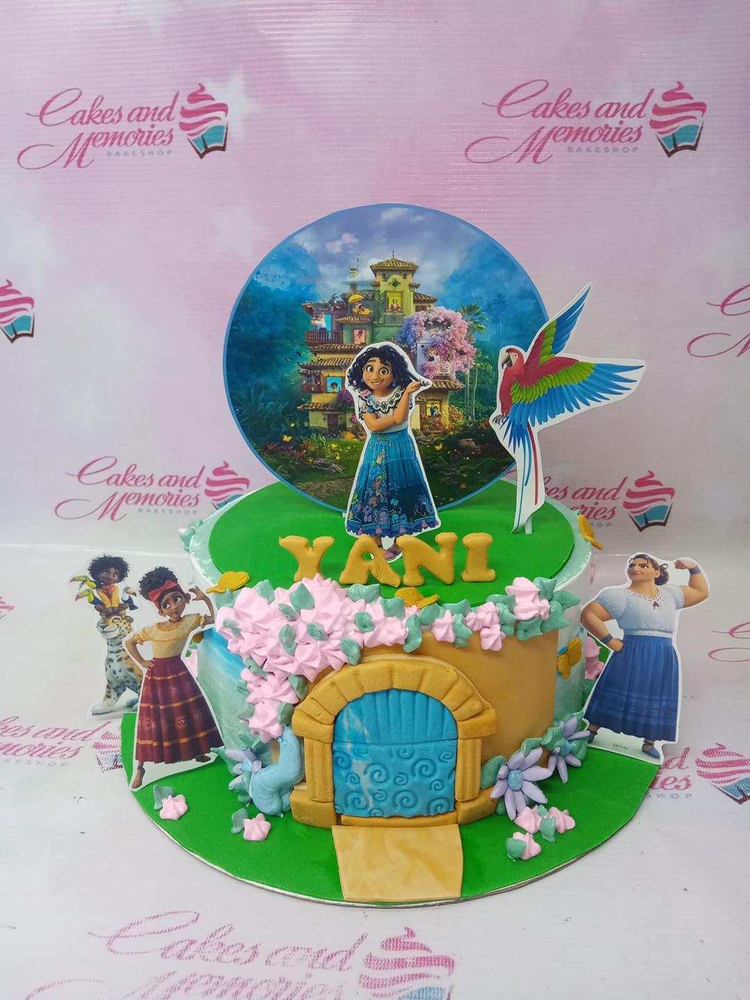 Encanto themed birthday cake with Mirabel and family printouts, a fondant door, and pink icing flowers on a green base.