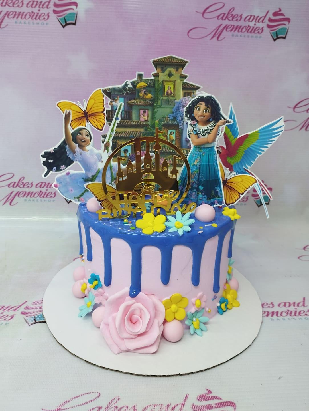 Encanto themed birthday cake with Mirabel and Isabela printouts, blue drip icing, and pink fondant roses.