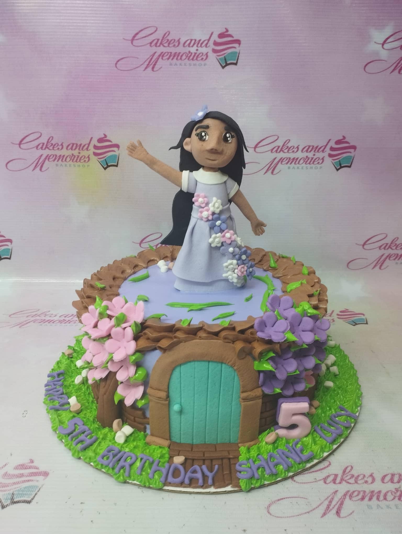 Encanto Isabela themed cake with a 3D fondant girl figure, purple and pink flowers, and a green door design on a grass-textured base.