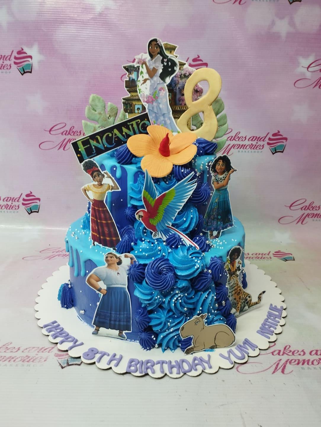 Two-tier blue Encanto themed birthday cake with character printouts, blue icing rosettes, and a large yellow hibiscus flower.
