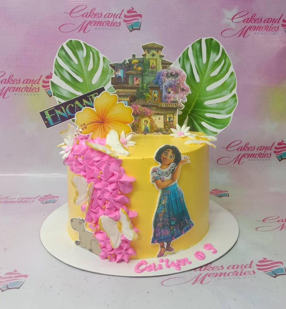 Yellow Encanto themed birthday cake with Mirabel printout, pink icing ruffles, and a large Casita house topper.