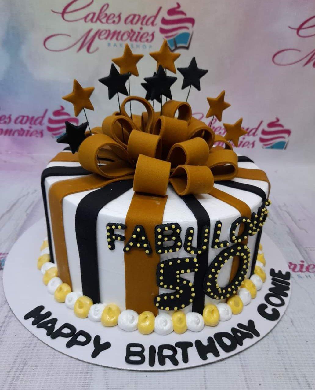 A white 1-tier birthday cake with black and gold vertical stripes, a large gold fondant bow, and black and gold star toppers.