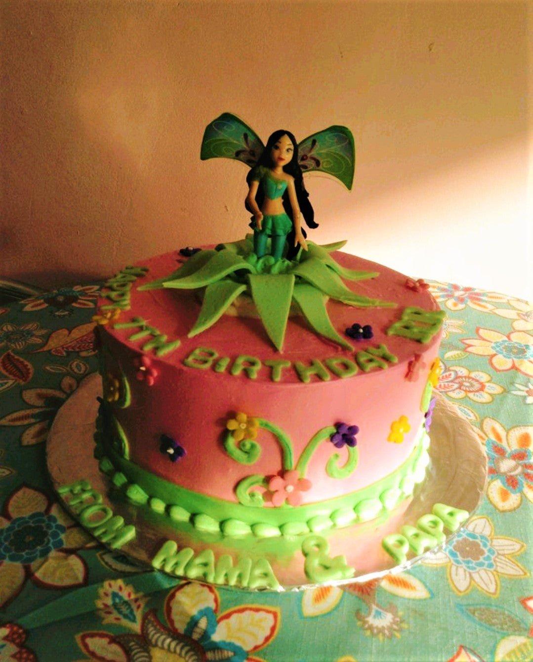 Pink fairy birthday cake with a 3D fairy figurine, green fondant leaves, and colorful icing flowers.
