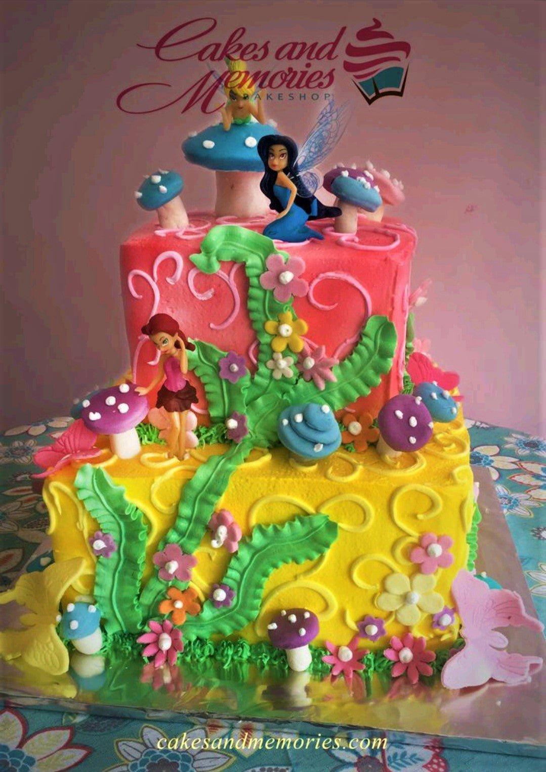 Two-tier square fairy garden cake with pink and yellow icing, fondant mushrooms, flowers, and fairy figurines.