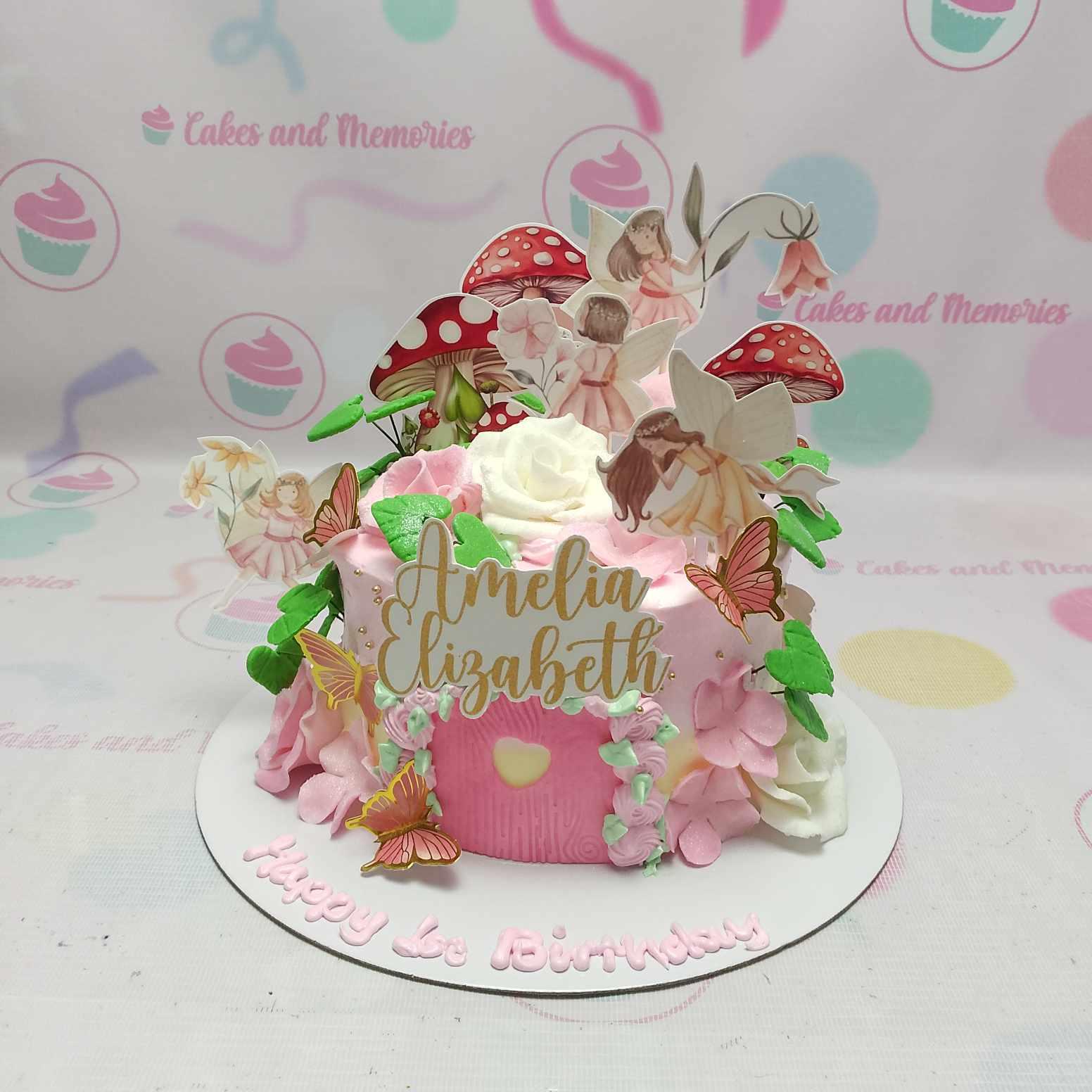 Pink fairy garden birthday cake with printed fairy and mushroom toppers, pink icing flowers, and a gold name topper.