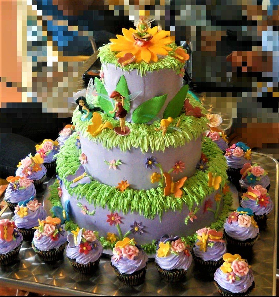 Three-tier purple fairy garden cake with green grass icing, fondant flowers, butterflies, and fairy figurines surrounded by matching cupcakes.
