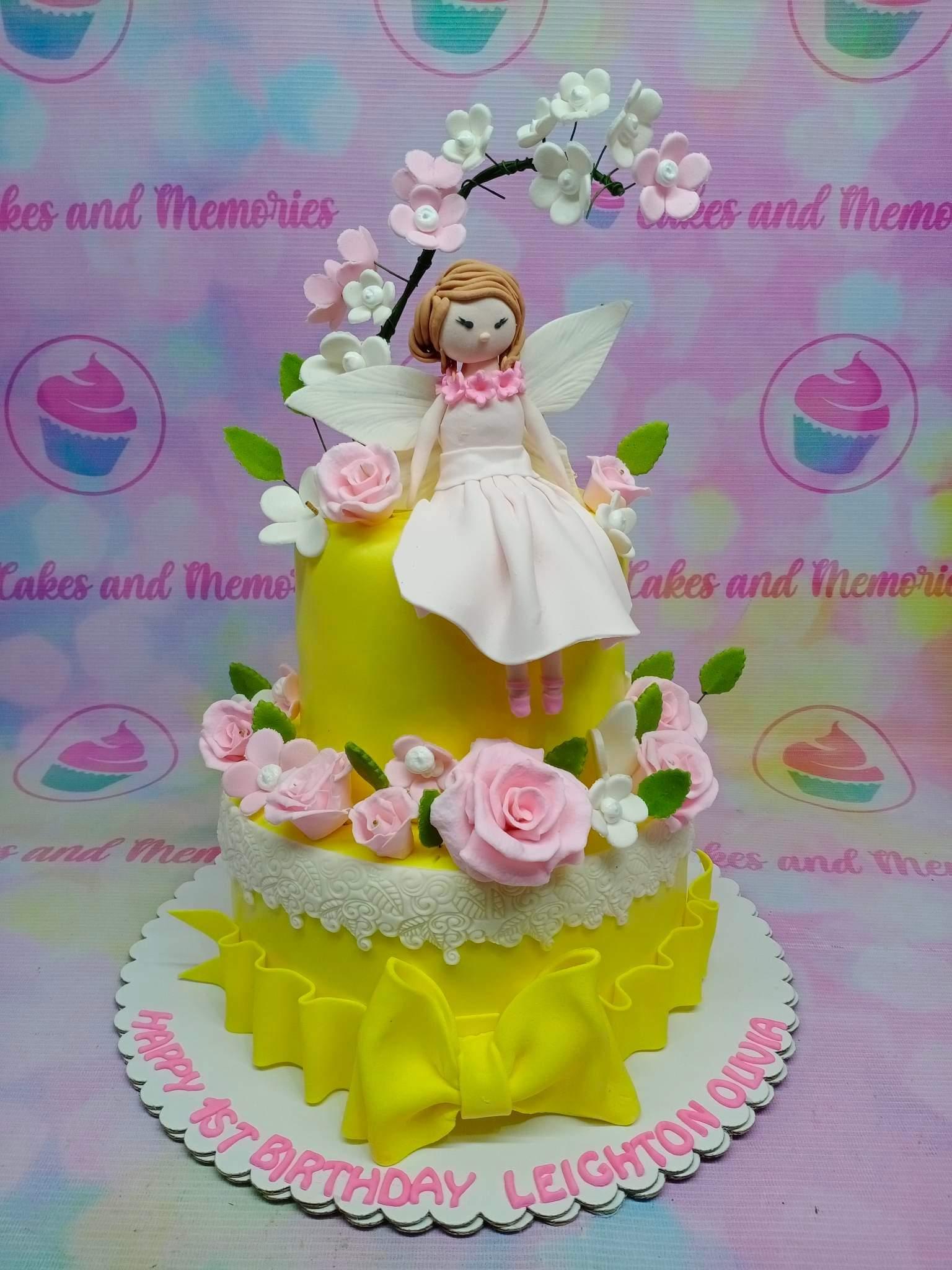 Yellow 2-tier fairy birthday cake with pink fondant roses, white blossoms, and a 3D fondant fairy topper.