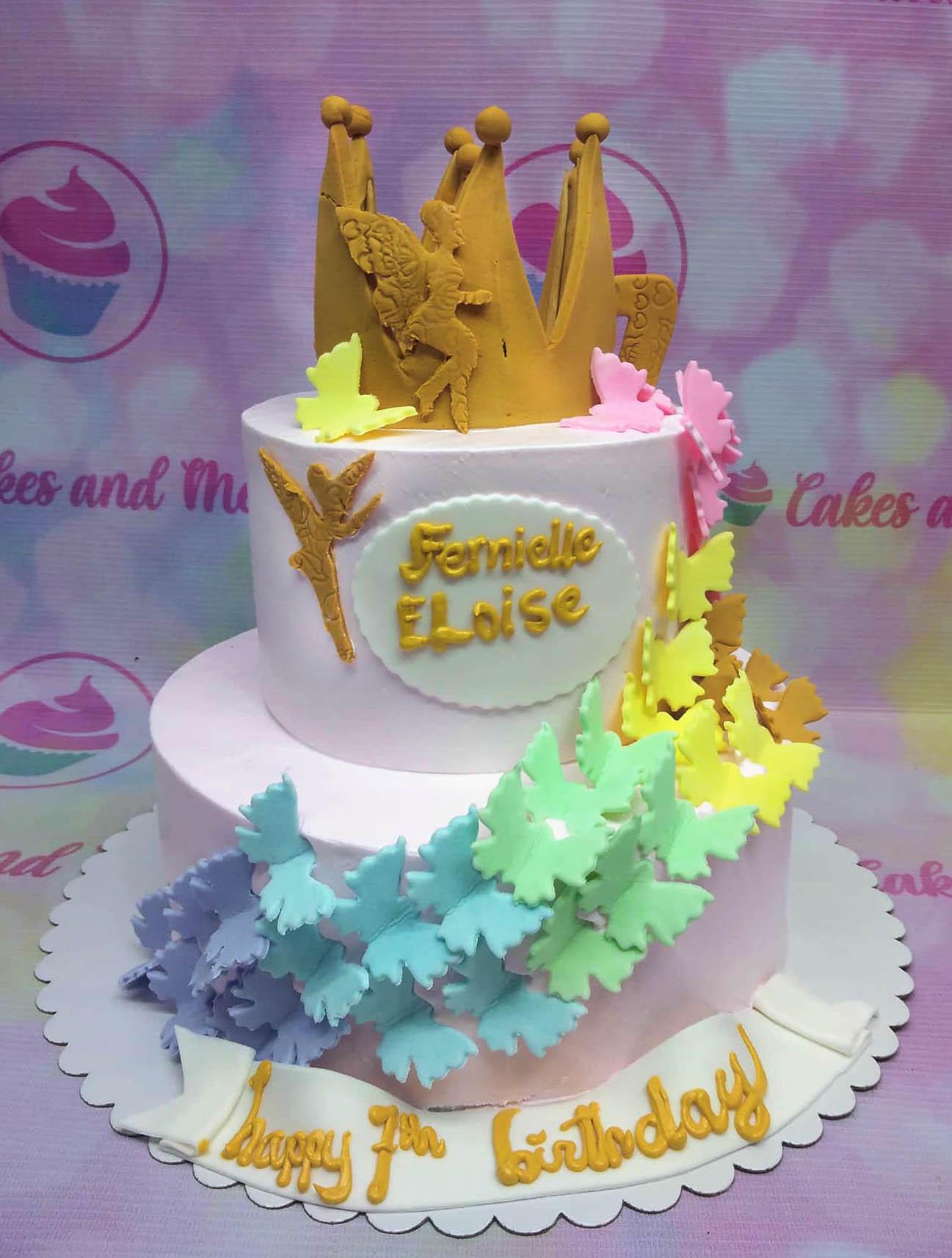 Two-tier light pink butterfly and fairy themed birthday cake with a gold crown topper and rainbow butterfly trail.