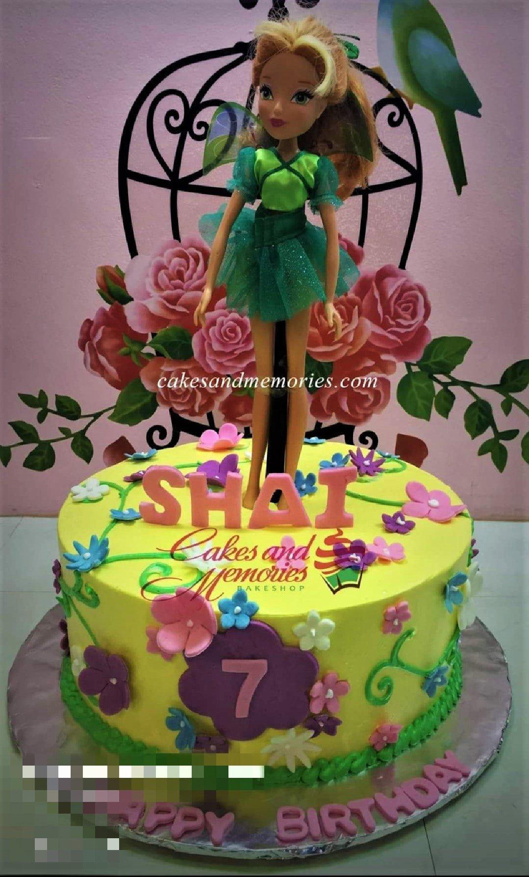 Yellow 1-tier birthday cake featuring a large fairy doll topper, pink gumpaste letters, and colorful floral decorations.