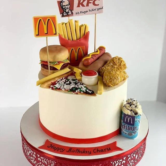 White 1-tier fast food themed birthday cake featuring fondant burger, fries, pizza, hot dog, fried chicken, and McFlurry toppers.