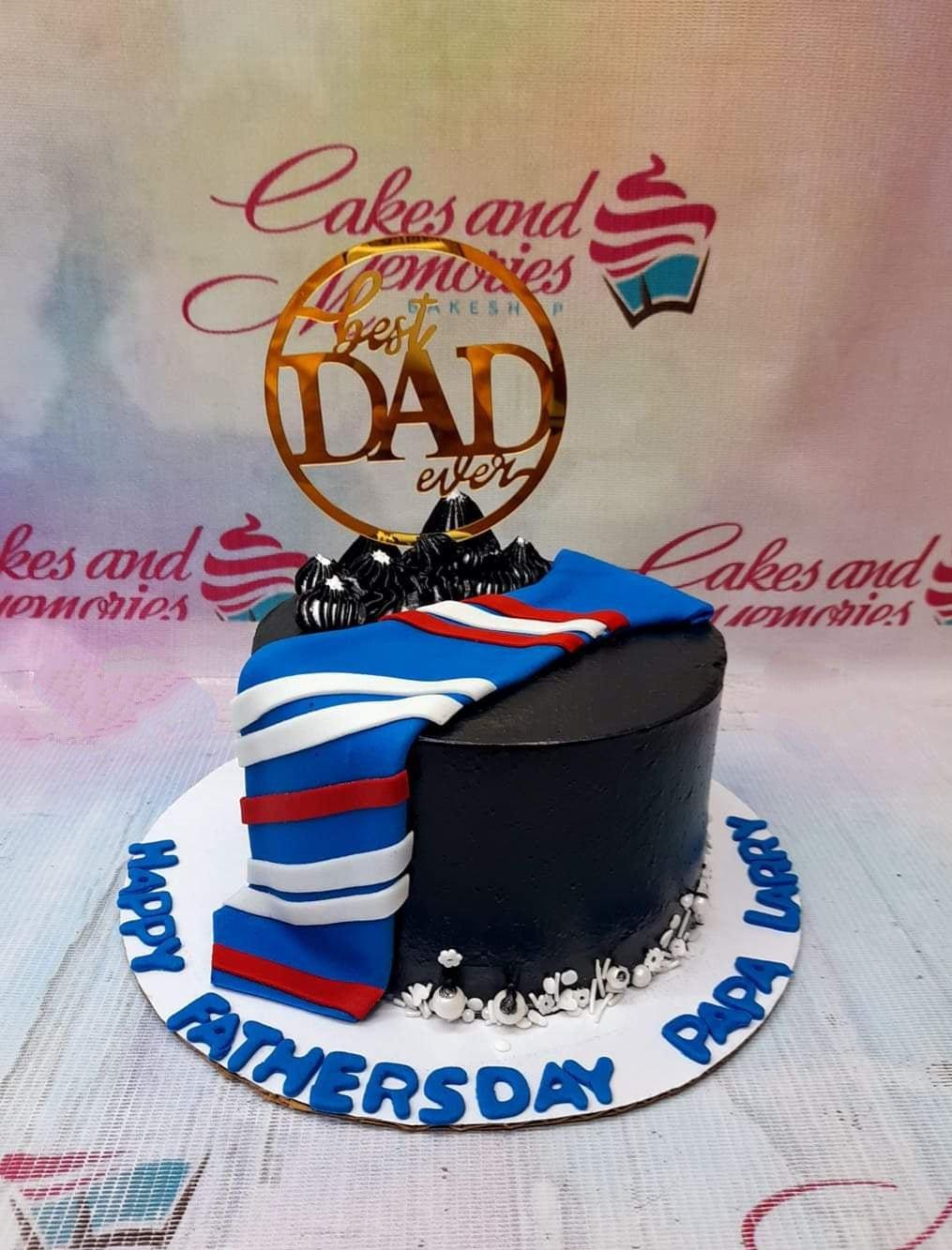Black Father's Day cake with a blue striped fondant tie, gold Best Dad Ever topper, and blue icing message on the base board.