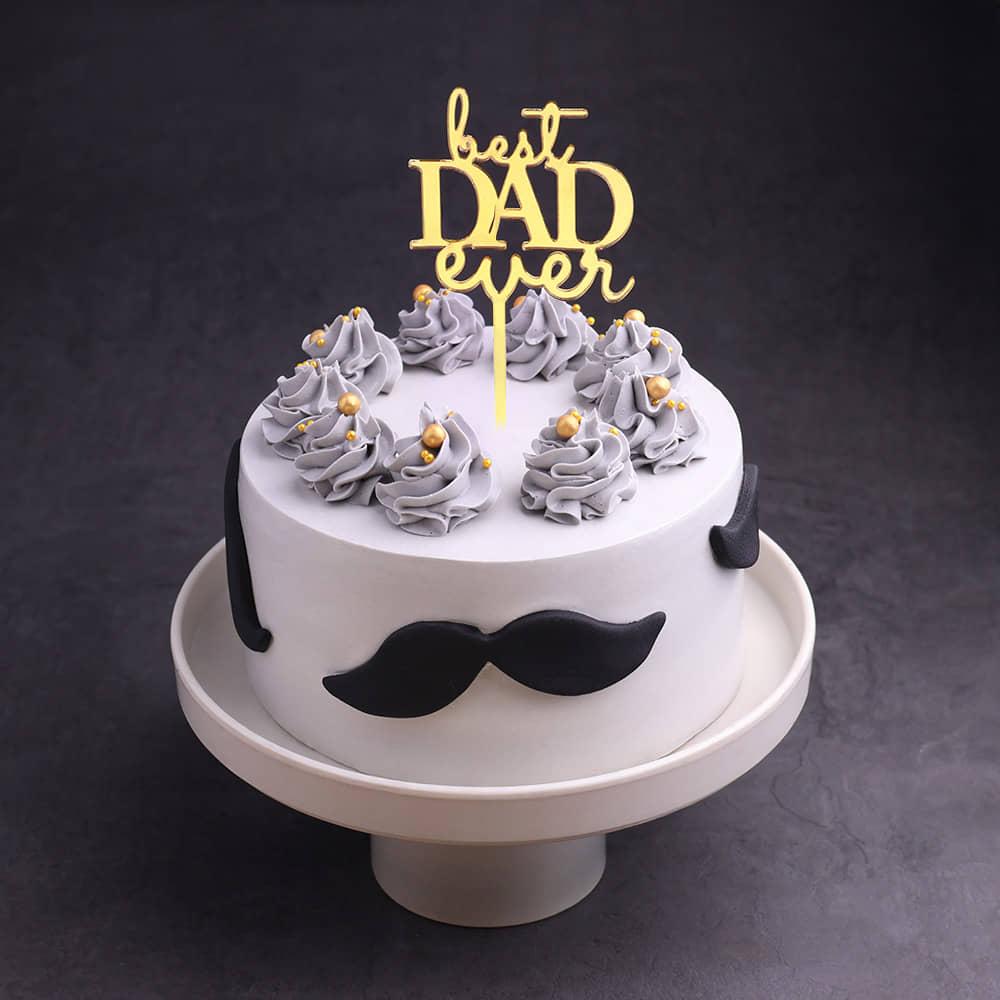 White minimalist cake for Father's Day featuring a gold Best Dad Ever topper, grey icing swirls with gold pearls, and black fondant mustaches.