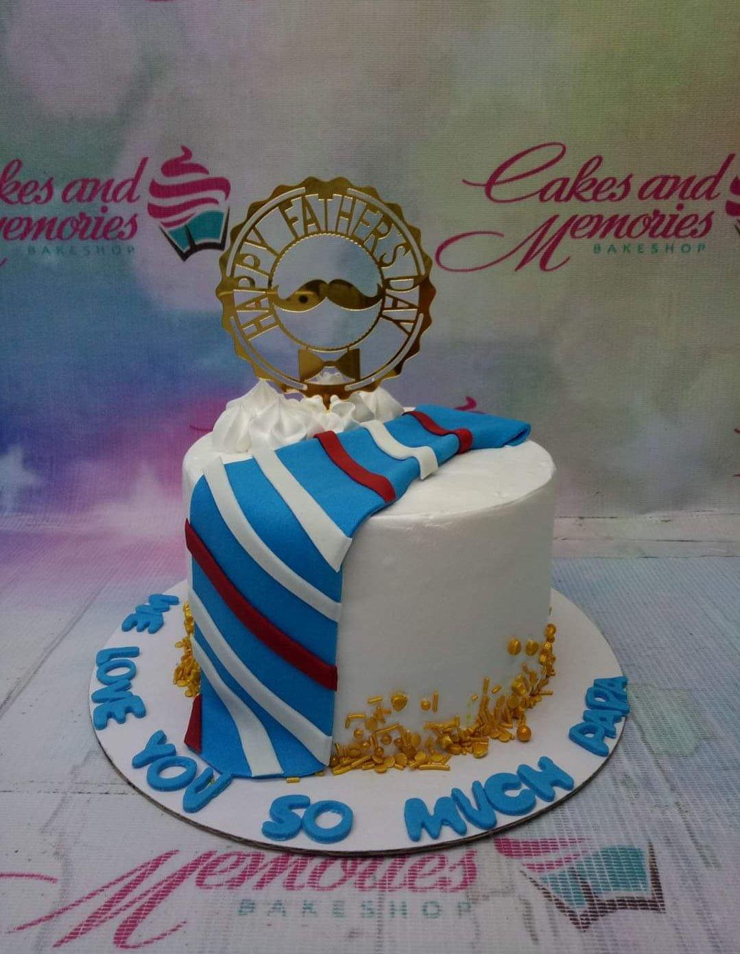 White Father's Day cake with a blue and white striped fondant tie, gold sprinkles, and a gold mustache topper.