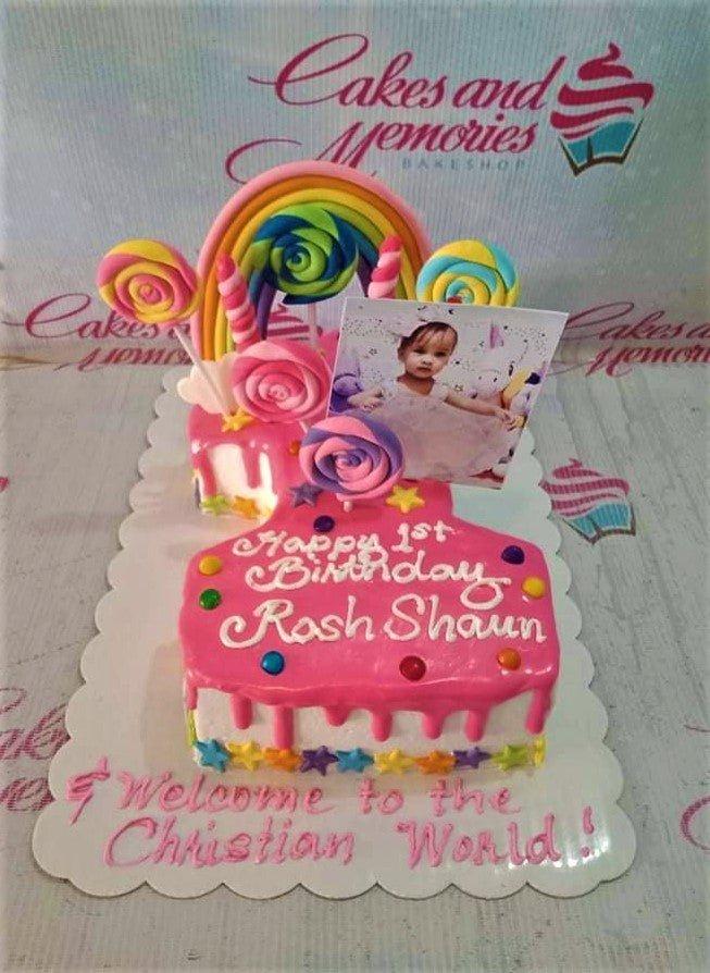 Pink number one shaped birthday cake with rainbow topper, lollipops, and a photo printout for a first birthday and christening.