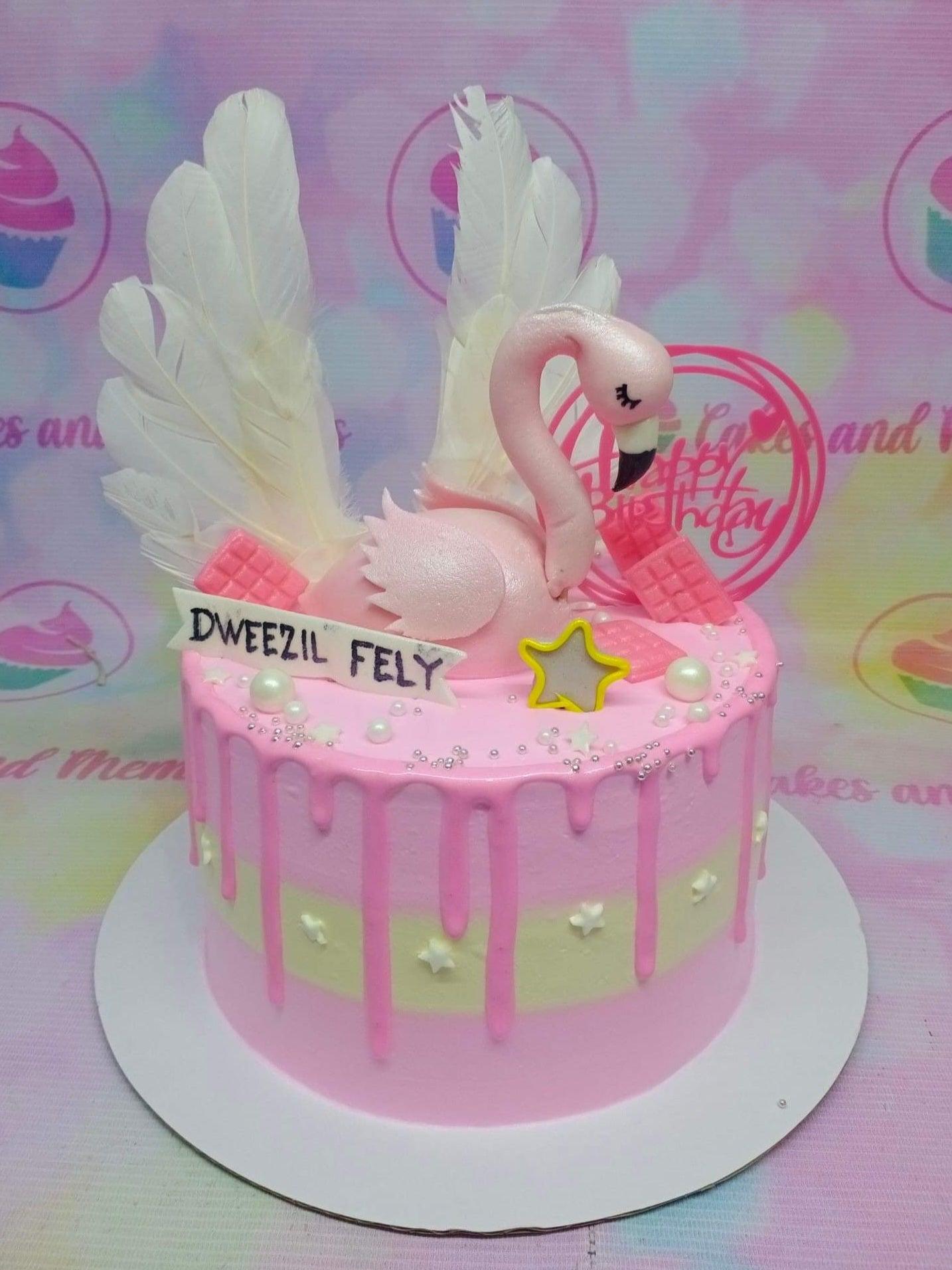 Pink flamingo birthday cake with white feather wings, pink chocolate bars, and silver dragees on a pink and yellow striped base.