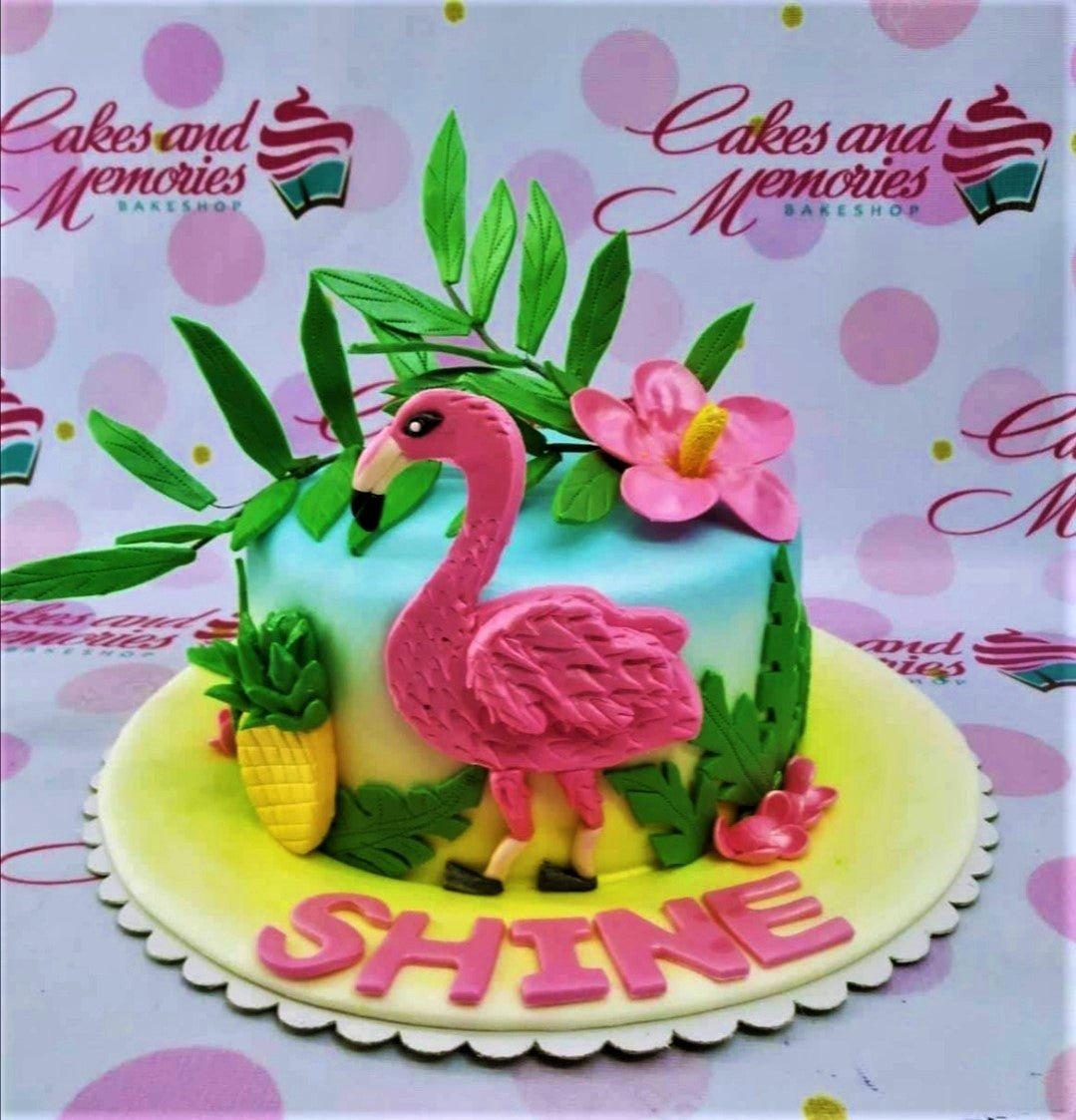 Tropical flamingo birthday cake with pink fondant flamingo, pineapple, hibiscus flower, and green leaves on a blue and yellow gradient base.